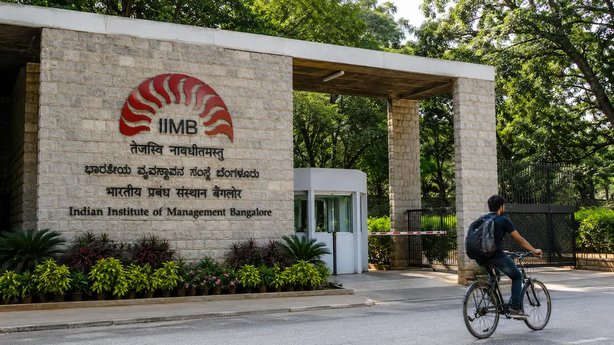 The stone brick entrance gate of the Indian Institute of Management Bangalore (IIMB) campus, featuring lush green trees and a cyclist passing by the main sign.