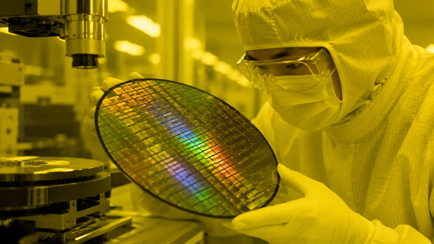 Close-up of an engineer inside a dust-free semiconductor cleanroom handling a silicon wafer under monochromatic light.