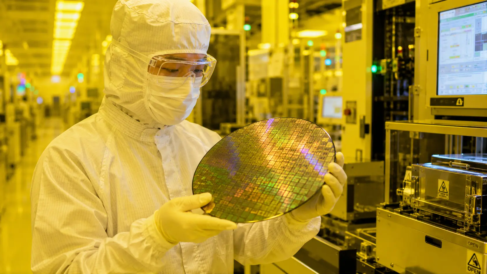 A semiconductor cleanroom technician wearing a full-body white sterile suit, protective mask, and goggles, carefully holding a reflective silicon microchip wafer under yellow cleanroom lighting inside a high-tech manufacturing fabrication facility.